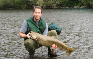 A photo of a seasoned angler smiling as he holds a king salmon while squatting in the water.