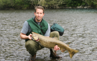 A photo of a seasoned angler smiling as he holds a king salmon while squatting in the water.